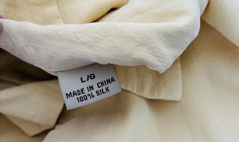 May include: Close-up of a cream-coloured silk garment with a label that reads "L/G MADE IN CHINA 100% SILK." The fabric has a subtle texture and sheen. The label is white with black text.