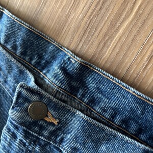 May include: Close-up of blue denim jeans. The image shows the waistband, button, and stitching details. The button is a dark metal color. The denim has a classic, worn look.
