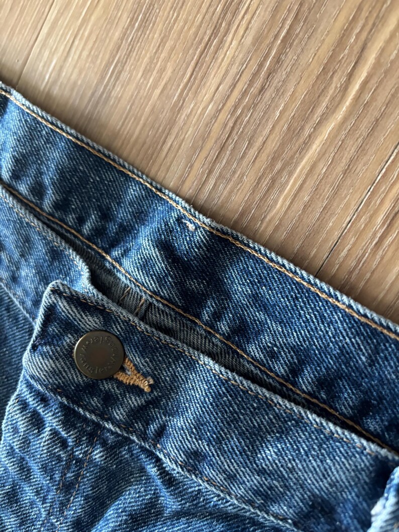 May include: Close-up of blue denim jeans. The image shows the waistband, button, and stitching details. The button is a dark metal color. The denim has a classic, worn look.
