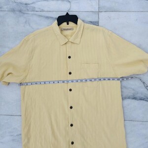 May include: A pale yellow short-sleeved shirt with a collar, chest pocket, and dark brown buttons. A measuring tape is laid across the shirt. The shirt has a brand label on the collar.