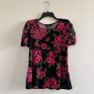 Peut inclure: Haut noir à manches courtes avec un motif floral rose vif. Le haut présente une encolure dégagée et des manches transparentes. Le motif floral comprend des fleurs roses et des feuilles vertes. Le haut est suspendu à un cintre noir sur fond blanc.