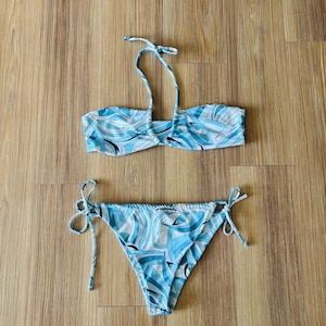 May include: A two-piece bikini set featuring a light blue, white, and black abstract graphic pattern. The top is a bandeau style with a thin halter neck strap. The bottom is a classic bikini cut with adjustable side ties on both hips.