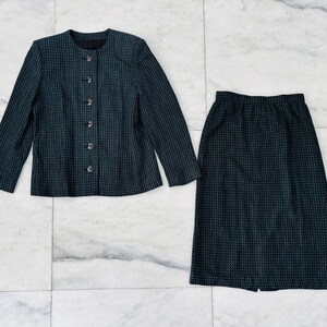 Puede incluir: Un conjunto de traje de dos piezas de color verde azulado oscuro. La chaqueta tiene cuello redondo y cierre de botones. La falda es hasta la rodilla. Ambas piezas tienen un patrón sutil y texturizado. El traje se muestra sobre una superficie de mármol.