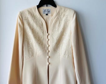 May include: A cream-colored blazer with a V-neck and long sleeves. The blazer has a floral embroidery design and a button closure. The label on the blazer reads "Le Suit".
