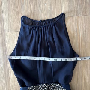May include: A sleeveless navy blue dress with a high, gathered neckline and a waistline embellished with silver-toned crystals. A white measuring tape is placed across the chest, showing a width measurement of approximately 16 inches.