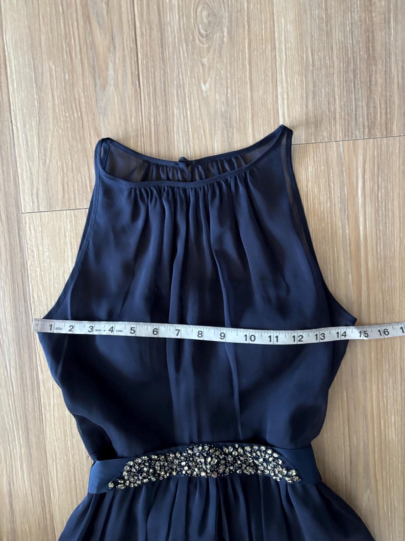 May include: A sleeveless navy blue dress with a high, gathered neckline and a waistline embellished with silver-toned crystals. A white measuring tape is placed across the chest, showing a width measurement of approximately 16 inches.