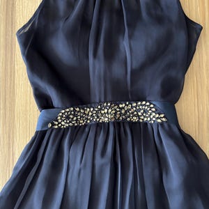 May include: A sleeveless navy blue dress featuring a high neckline and a gathered bodice. The waist is accented with a horizontal band embellished with dark, faceted crystals arranged in an intricate, ornate pattern above a pleated skirt.