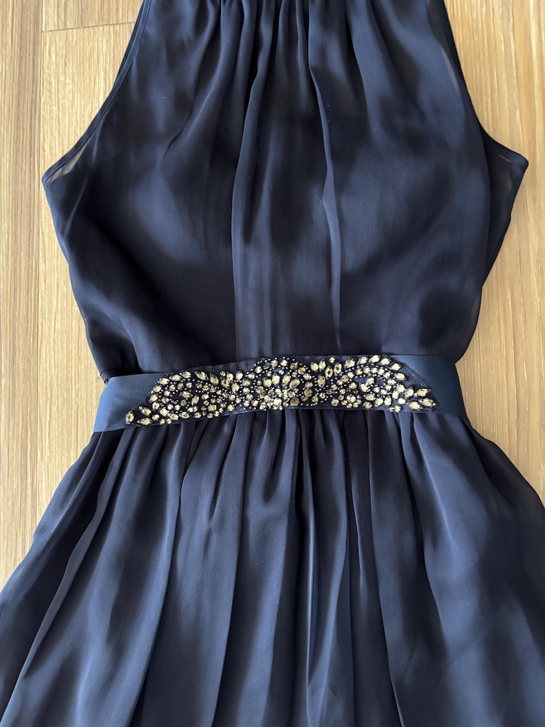 May include: A sleeveless navy blue dress featuring a high neckline and a gathered bodice. The waist is accented with a horizontal band embellished with dark, faceted crystals arranged in an intricate, ornate pattern above a pleated skirt.