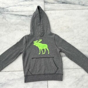 May include: A gray hooded sweatshirt with a kangaroo pocket and a neon green moose graphic on the front. The sweatshirt has long sleeves and a drawstring hood. The material appears to be a soft, comfortable fabric.