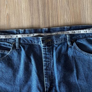 May include: Close-up of blue denim jeans with a measuring tape across the waistband. The waist measures approximately 24 inches. The jeans have classic five-pocket styling.