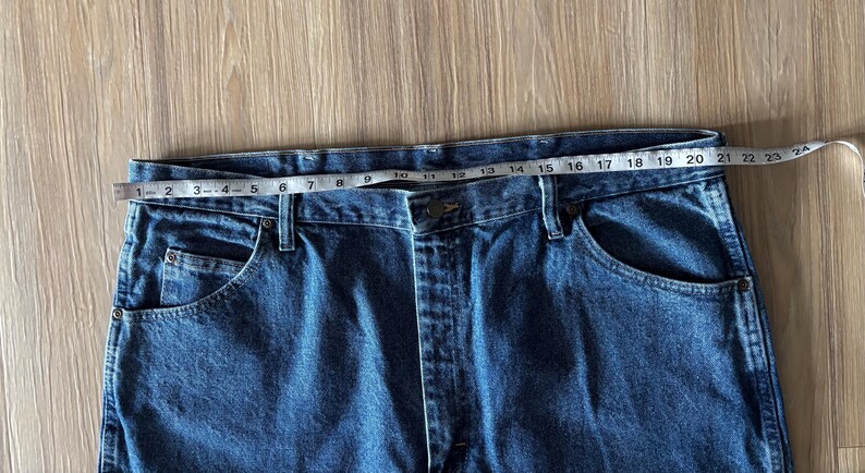 May include: Close-up of blue denim jeans with a measuring tape across the waistband. The waist measures approximately 24 inches. The jeans have classic five-pocket styling.