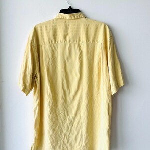 May include: A pale yellow short-sleeved shirt on a black hanger against a white wall. The shirt features a collared neckline and a button-down design. The back of the shirt is visible, showing a yoke and a slightly textured fabric.