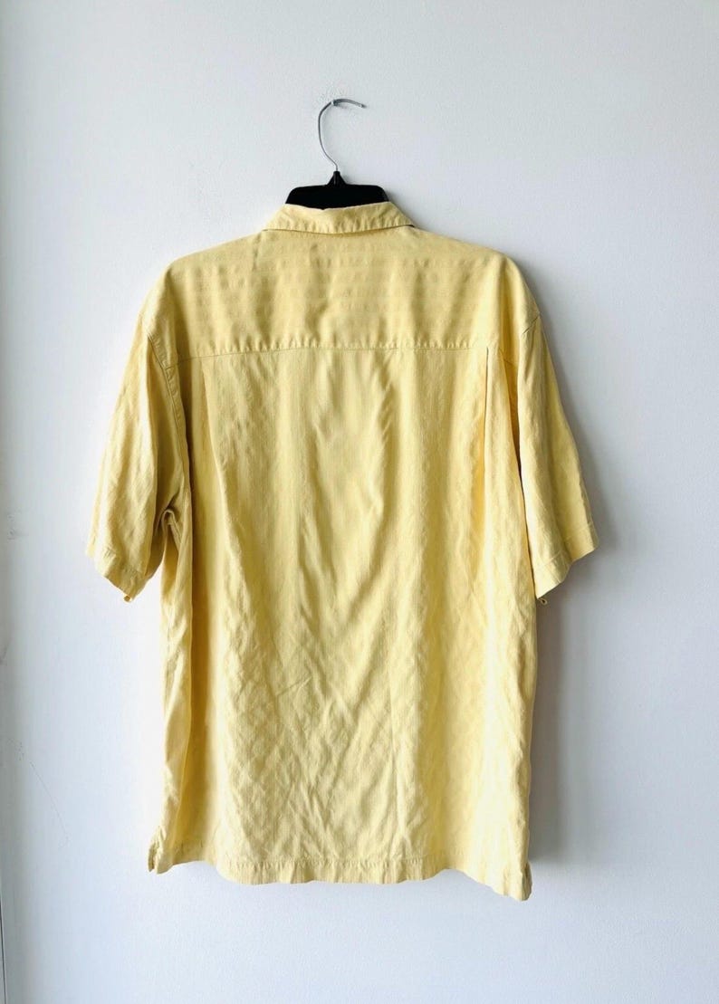 May include: A pale yellow short-sleeved shirt on a black hanger against a white wall. The shirt features a collared neckline and a button-down design. The back of the shirt is visible, showing a yoke and a slightly textured fabric.
