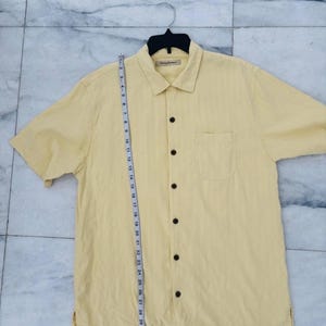 May include: A pale yellow short-sleeved shirt with a button-down front and a chest pocket. The shirt is hanging on a black hanger. A measuring tape is placed next to the shirt, showing measurements in centimetres.