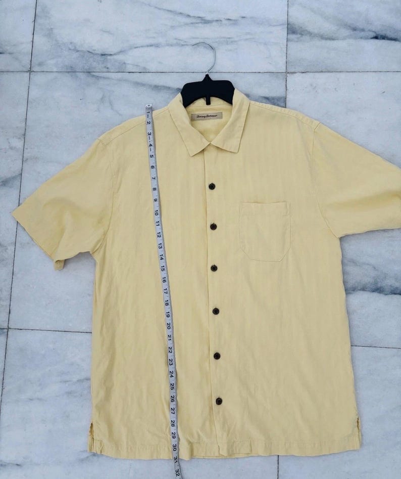 May include: A pale yellow short-sleeved shirt with a button-down front and a chest pocket. The shirt is hanging on a black hanger. A measuring tape is placed next to the shirt, showing measurements in centimetres.