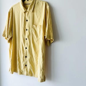 May include: A pale yellow short-sleeved shirt with a single chest pocket. The shirt is suspended from a black hanger against a white wall. The shirt has a collar and dark brown buttons.