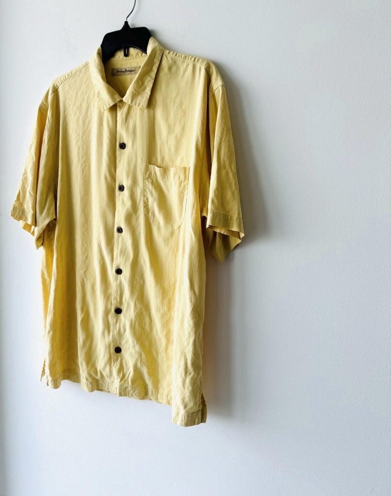 May include: A pale yellow short-sleeved shirt with a single chest pocket. The shirt is suspended from a black hanger against a white wall. The shirt has a collar and dark brown buttons.