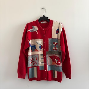 Könnte beinhalten: Roter Cardigan mit Knopfleiste und langen Ärmeln. Der Pullover hat ein Patchwork-Design mit gestickten Bildern in Blau, Creme und Grau. Die Knöpfe sind silberfarben. Das Kleidungsstück hängt an einem schwarzen Kleiderbügel.