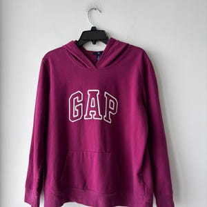 May include: A magenta hoodie with the word "GAP" in white block letters across the chest. The hoodie has a kangaroo pocket and a drawstring hood. The sweatshirt is hanging on a black hanger against a white wall.