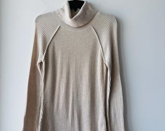 May include: A beige ribbed knit turtleneck sweater hanging on a black hanger against a white wall. The long-sleeved sweater has a high, folded collar and vertical ribbing throughout the fabric. The sweater's neutral color makes it a versatile wardrobe staple.