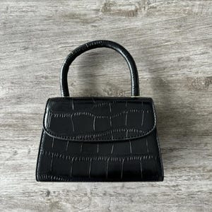 Könnte beinhalten: Schwarze Handtasche mit einem strukturierten, krokodilartigen Muster. Die kleine Geldbörse hat einen gebogenen Griff und einen Klappenverschluss. Die Tasche steht auf einer hellgrauen Holzoberfläche.