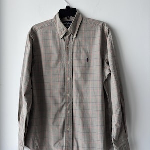 May include: A long-sleeved button-down shirt in a plaid pattern of brown, red, and navy blue. The shirt has a button-down collar and a small purple logo on the chest. The shirt is hanging on a black hanger.