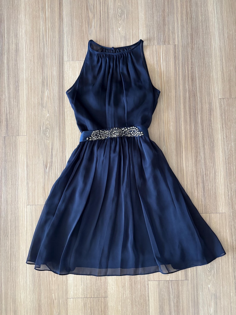 May include: A navy blue sleeveless dress featuring a high halter neckline with gathered fabric details. The waist is accented by a horizontal band embellished with clear, sparkling crystals and beads, leading into a pleated, knee-length skirt.