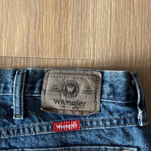 May include: Close-up of blue denim Wrangler jeans. The jeans feature a brown leather patch with the Wrangler logo and text. A red tag with the Wrangler logo is sewn on the back pocket. The jeans are made of durable denim.