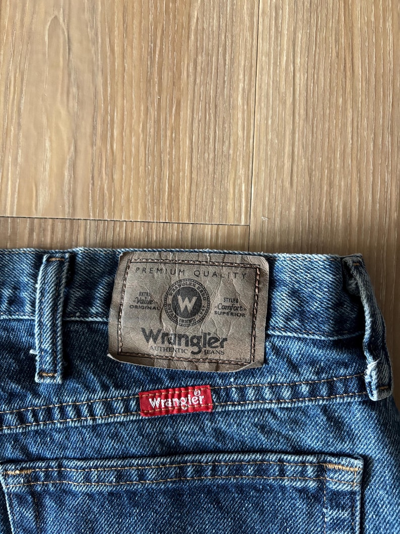May include: Close-up of blue denim Wrangler jeans. The jeans feature a brown leather patch with the Wrangler logo and text. A red tag with the Wrangler logo is sewn on the back pocket. The jeans are made of durable denim.