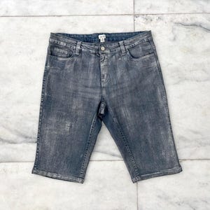 May include: A pair of gray denim Bermuda shorts. The shorts have a button and zipper closure, front and back pockets, and a faded, distressed appearance. The shorts are laid flat on a marble surface.