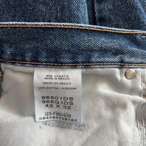 May include: Close-up of blue denim jeans. The jeans have a classic five-pocket design and a button and zipper closure. The tag reads "MADE IN MEXICO" and "100% COTTON". The size is 42 x 32.