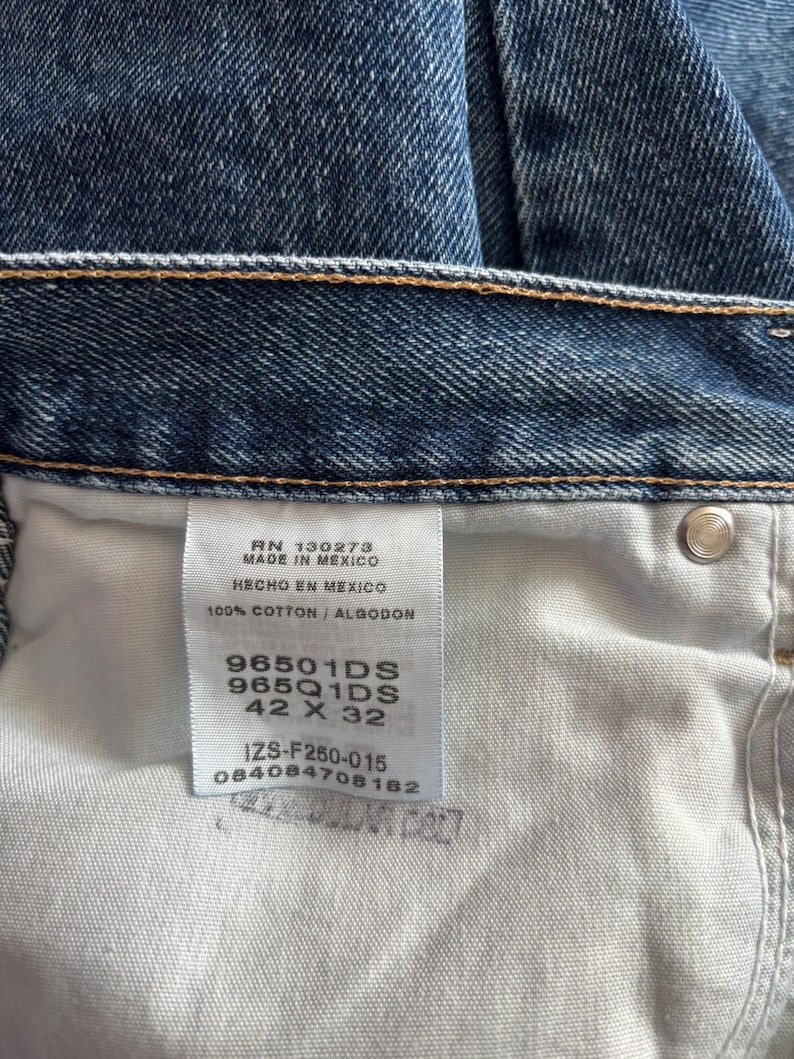 May include: Close-up of blue denim jeans. The jeans have a classic five-pocket design and a button and zipper closure. The tag reads "MADE IN MEXICO" and "100% COTTON". The size is 42 x 32.
