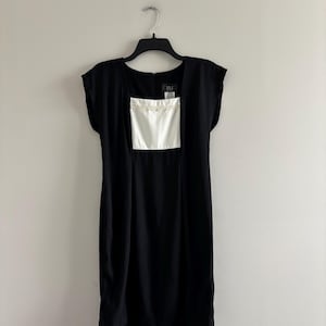 Puede incluir: Un vestido negro con un panel cuadrado blanco en el corpiño, colgado de una percha negra. El vestido tiene un escote cuadrado y mangas cortas. El panel blanco está adornado con un delicado diseño de cuentas. El vestido es de un estilo clásico y elegante.