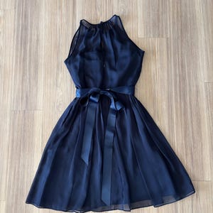 May include: A sleeveless navy blue dress featuring a high halter neckline with a sheer overlay, a fitted bodice, and a pleated skirt. The waist is accented with a matching navy blue satin ribbon tied into a bow at the center front.