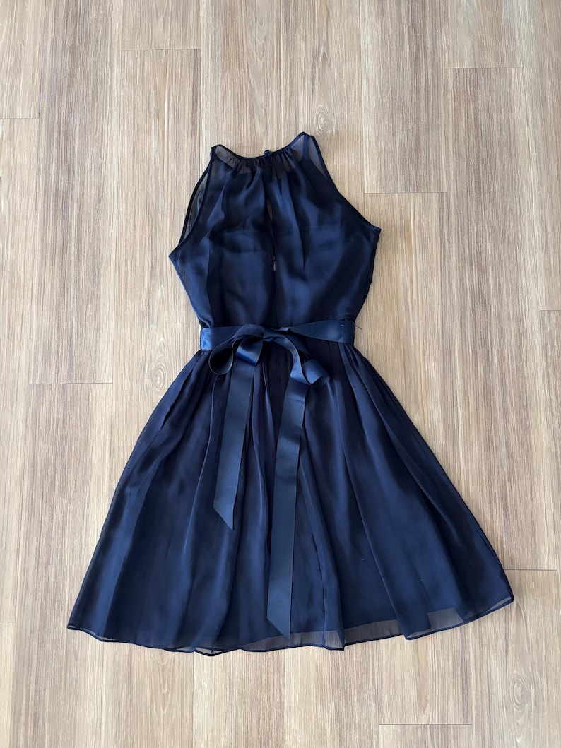 May include: A sleeveless navy blue dress featuring a high halter neckline with a sheer overlay, a fitted bodice, and a pleated skirt. The waist is accented with a matching navy blue satin ribbon tied into a bow at the center front.