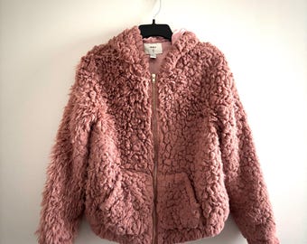 May include: A dusty rose-colored hooded jacket with a zip-up front and two front pockets. The jacket is made of a soft, textured material and is hanging on a black hanger against a white wall.