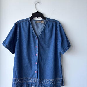 Könnte beinhalten: Kurzärmeliges Jeanshemd mit V-Ausschnitt und rosa Knöpfen. Das Hemd hat einen dekorativen, gestickten Saum. Das Hemd hängt an einem schwarzen Kleiderbügel vor einer weißen Wand.