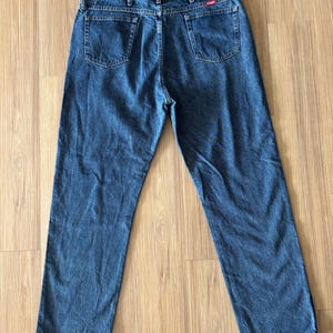 May include: Blue denim jeans with a classic five-pocket design. The jeans have a straight leg cut and a Wrangler brand patch on the back. The jeans are laid flat on a wooden surface.