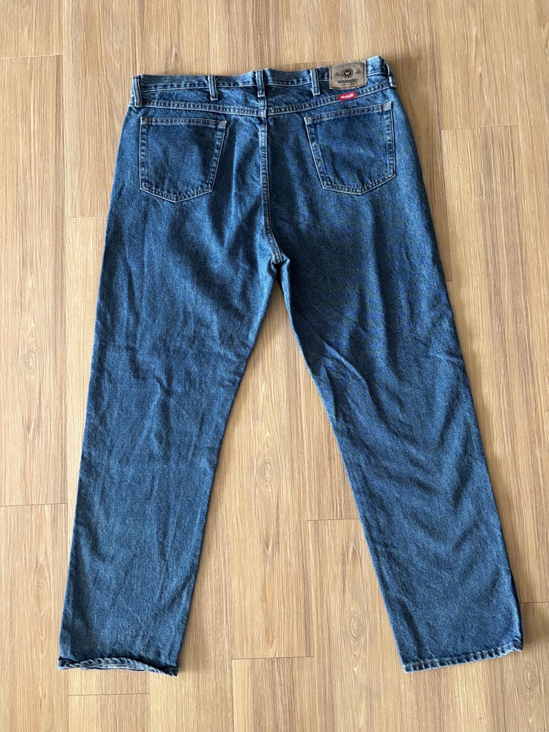 May include: Blue denim jeans with a classic five-pocket design. The jeans have a straight leg cut and a Wrangler brand patch on the back. The jeans are laid flat on a wooden surface.