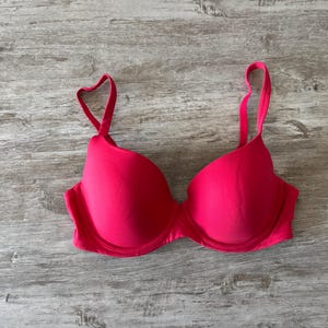May include: A vibrant, hot pink bra with adjustable straps. The bra features molded cups and underwire support. The straps are a matching pink and are attached to the cups with a heart-shaped detail. The bra is made of a smooth, solid fabric.
