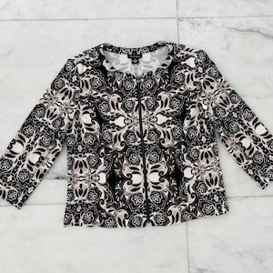 May include: A black, white, and tan patterned jacket with a zipper closure. The jacket features a floral design and long sleeves. The garment is laid flat on a marble surface.