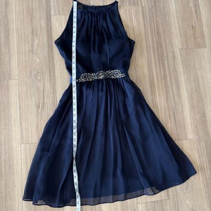 May include: A navy blue sleeveless dress with a high neckline and a pleated skirt. A horizontal band at the waist is embellished with silver-toned beads and crystals. A white measuring tape is placed vertically, showing a length of 36 inches.