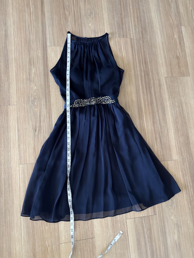 May include: A navy blue sleeveless dress with a high neckline and a pleated skirt. A horizontal band at the waist is embellished with silver-toned beads and crystals. A white measuring tape is placed vertically, showing a length of 36 inches.