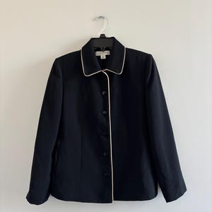 Pode incluir: Um blazer preto com acabamento creme ao redor da gola e das bordas frontais. O blazer tem fechamento por botões e mangas compridas. A peça está pendurada em um cabide preto contra uma parede branca.