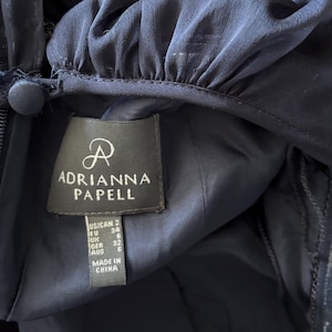 May include: A dark navy blue garment featuring a gathered neckline with a fabric-covered button closure and a side zipper. A rectangular label displays the brand name Adrianna Papell, size US 2, EU 34, UK 6, GER 32, AUS 6, and Made in China.