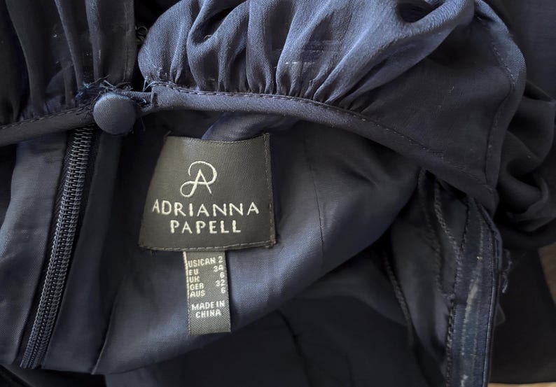 May include: A dark navy blue garment featuring a gathered neckline with a fabric-covered button closure and a side zipper. A rectangular label displays the brand name Adrianna Papell, size US 2, EU 34, UK 6, GER 32, AUS 6, and Made in China.