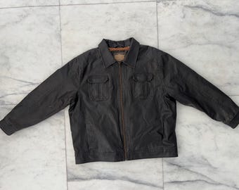 Puede incluir: Chaqueta gris oscuro con cuello, dos bolsillos en el pecho y cremallera completa. La chaqueta tiene mangas largas y forro interior marrón. La chaqueta está tendida sobre una superficie de mármol.