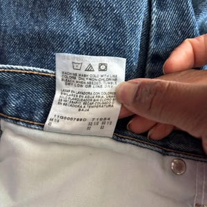 May include: Close-up of blue denim jeans with a white tag. The tag displays washing instructions and size information. The jeans are a classic style, with a button and a metal rivet visible.