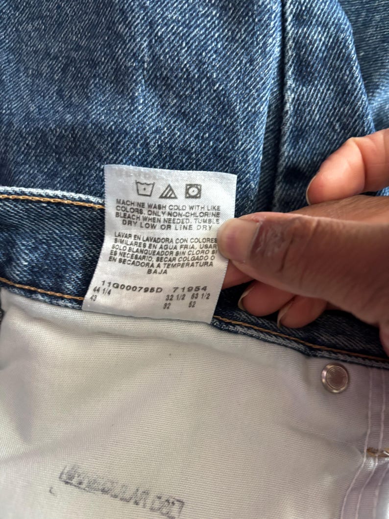 May include: Close-up of blue denim jeans with a white tag. The tag displays washing instructions and size information. The jeans are a classic style, with a button and a metal rivet visible.
