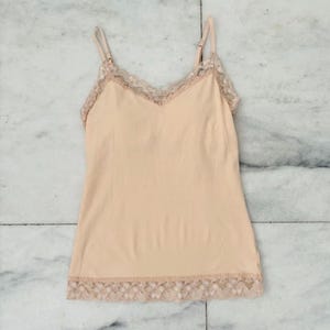May include: A peach-coloured camisole with delicate spaghetti straps and a V-neckline. The top is trimmed with a light brown lace detail along the neckline and hem. The camisole is made of a soft, lightweight material.